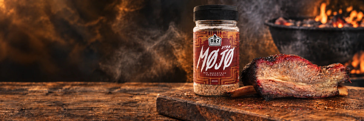 Campaign artwork showing Xtra Mojo seasoning beside smoked bone-in beef ribs on a timber board, lit by a charcoal barbecue